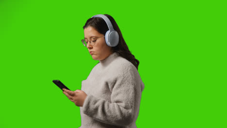Junge-Frau-Mit-Kabellosen-Kopfhörern,-Die-Musik-Vom-Mobiltelefon-Vor-Grünem-Studiobildschirm-Streamen-1